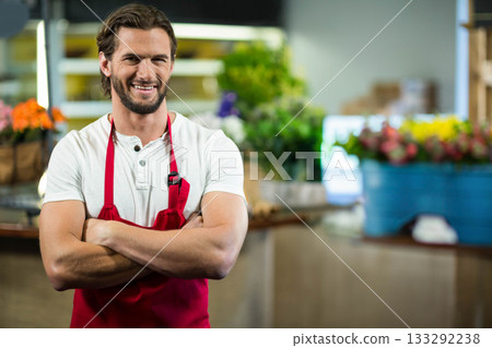 Male florist wearing red apron crossing arms at flower shop near buckets of blooms, copy space 133292238