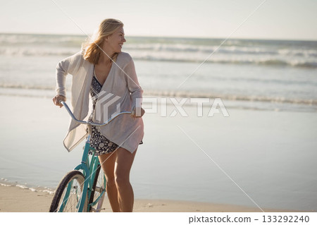 Blond female walking on wet beach pushing turquoise cruiser bicycle wearing light cardigan 133292240
