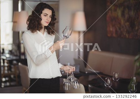 Female server wearing white blouse examining wine glasses under table lamp light in restaurant 133292246