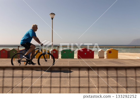 Senior man riding bicycle along seaside path past railing, beach huts and lamppost, copy space 133292252