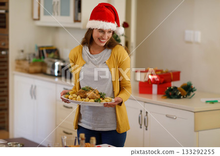 Woman holding platter of roast chicken and potatoes and wearing Santa hat with gifts in kitchen Woman holding platter of roast chicken and potatoes and wearing Santa hat with gifts in kitchen 133292255