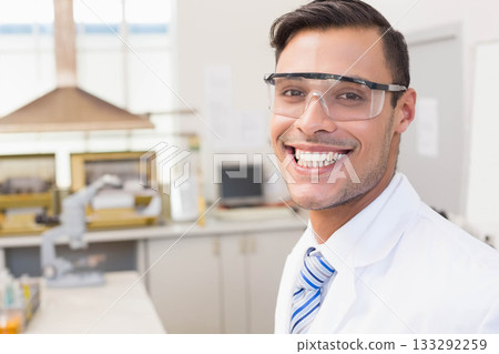 Man wearing lab coat, striped tie and safety goggles examining sample under microscope on lab bench Man wearing lab coat, striped tie and safety goggles examining sample under microscope on lab bench 133292259