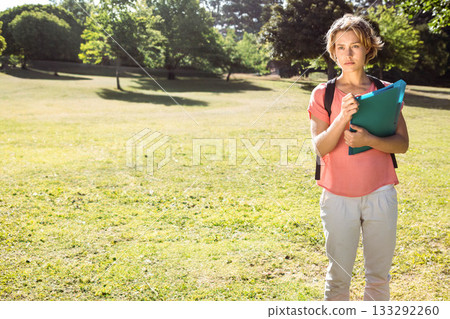 Woman student standing in sunlit park on grassy lawn holding teal folder and backpack, copy space 133292260