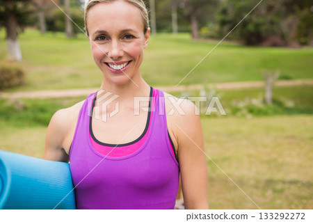 Mid adult woman holding rolled blue yoga mat wearing pink tank top in grassy park 133292272