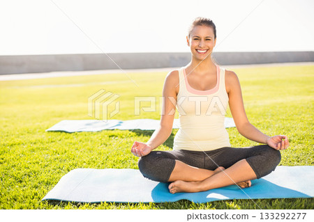 Mid adult woman meditating on blue yoga mat on grass under sunshine with pale mat behind 133292277