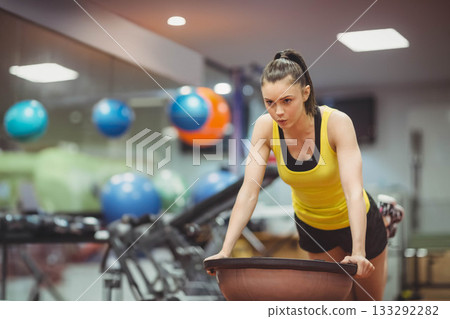 Female leaning forward on BOSU balance trainer in gym near wall-mounted stability balls, copy space 133292282