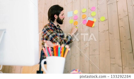 Man placing colorful round and square sticky notes on wooden accent wall in modern office workspace 133292284
