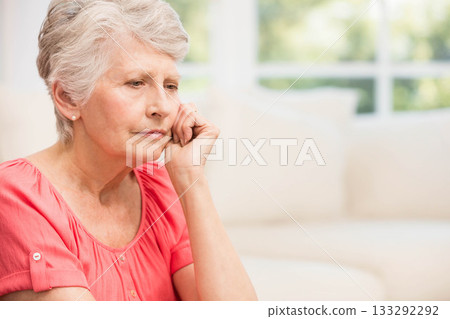 Senior woman gazing downward while resting on sofa by window in living room, copy space 133292292