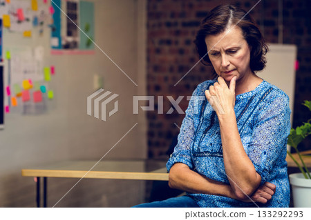 Senior female wearing blue blouse and jeans thinking at office table with planning board and plant Senior female wearing blue blouse and jeans thinking at office table with planning board and plant 133292293