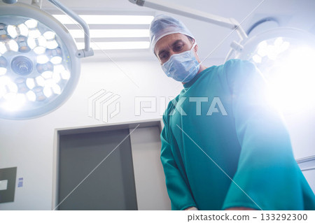 Male surgeon standing in scrubs under lamps in operating room, inspecting surgical field 133292300
