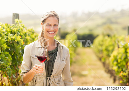 Woman standing in vineyard corridor wearing trench jacket holding red wine glass with sunlit vines Woman standing in vineyard corridor wearing trench jacket holding red wine glass with sunlit vines 133292302