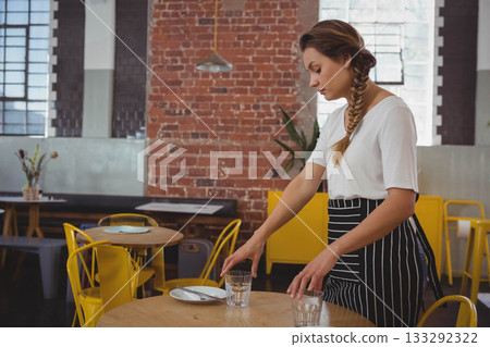 Female server wearing apron arranging clear glasses, plate, cutlery on cafe table, copy space Female server wearing apron arranging clear glasses, plate, cutlery on cafe table, copy space 133292322