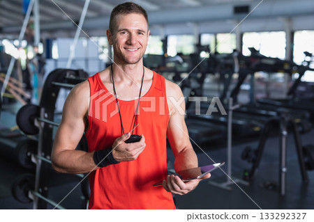 Mid adult trainer wearing athletic top holding stopwatch tablet in gym near weight benches, mirrors 133292327