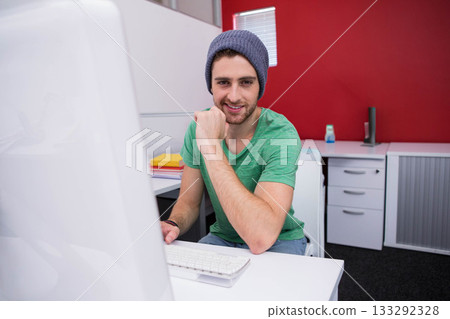 Male wearing green t-shirt and beanie sitting at desk in cubicle, leaning on fist beside notebooks Male wearing green t-shirt and beanie sitting at desk in cubicle, leaning on fist beside notebooks 133292328