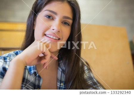 Woman wearing plaid shirt sitting on wooden bench in classroom, smiling gently resting chin on hand 133292340