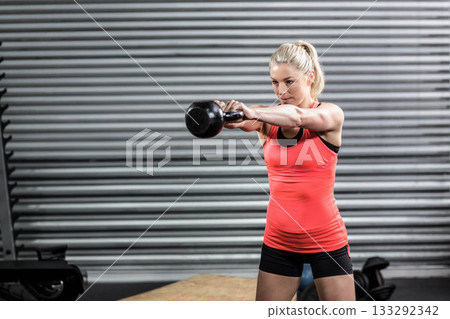 Mid adult woman wearing red top and black shorts performing kettlebell swing at wooden gym platform Mid adult woman wearing red top and black shorts performing kettlebell swing at wooden gym platform 133292342