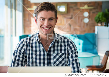 Male smiling while working on laptop in bright lounge area with turquoise sofa and plant 133292357