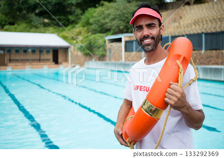 Male lifeguard with red visor holding orange rescue buoy with rope beside pool lanes, copy space 133292369