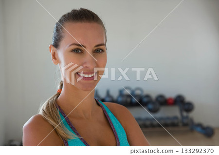 Female standing arms crossed in fitness studio next to kettlebell dumbbell rack, copy space 133292370