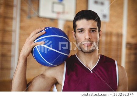Male basketball player in jersey holding blue basketball on shoulder inside gym under mounted hoop 133292374