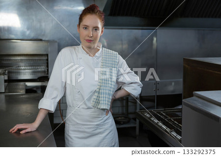 Female chef wearing chef jacket and checkered towel leaning on counter in kitchen with ovens 133292375