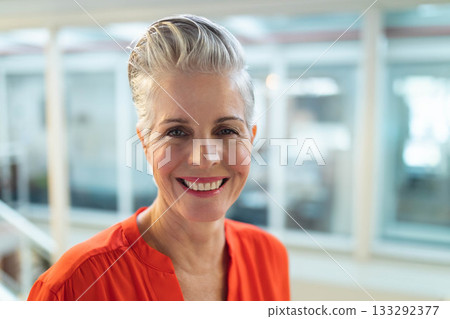 Senior woman wearing bright orange blouse, smiling and standing in office corridor with glass walls 133292377