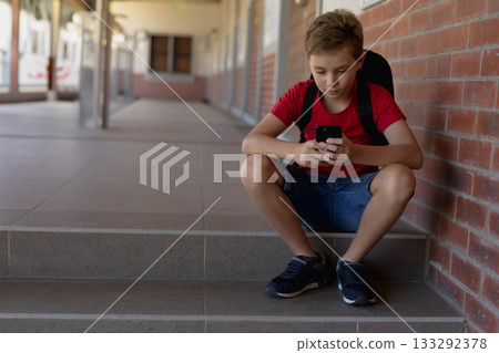 Child boy sitting on tiled steps under covered walkway using smartphone with backpack, copy space 133292378