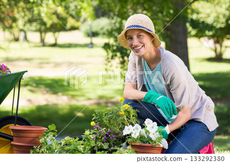 Woman kneeling in sunny garden wearing straw hat planting potted flowers by wheelbarrow, copy space 133292380
