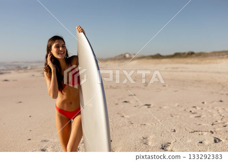 Woman wearing red swimsuit standing on sandy beach holding surfboard near water with pebbles 133292383