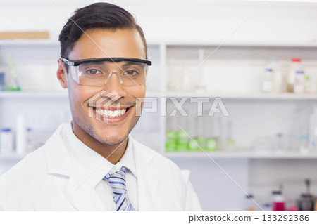 Smiling male scientist in safety goggles, lab coat, striped tie posing in lab with green solution Smiling male scientist in safety goggles, lab coat, striped tie posing in lab with green solution 133292386