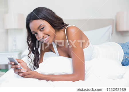 African American woman lying on bed propping on elbows and holding smartphone on white bedding African American woman lying on bed propping on elbows and holding smartphone on white bedding 133292389