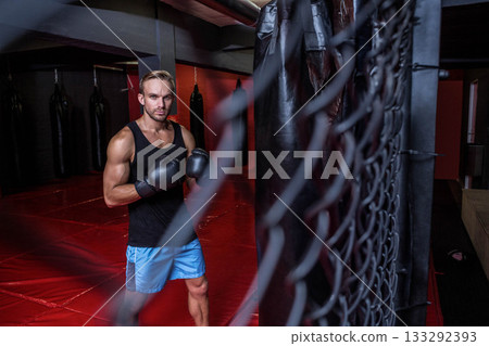 Male athlete training punches against heavy bag on red padded floor in gym with boxing gloves 133292393