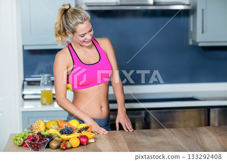Woman in twenties wearing sportswear leaning on home kitchen counter with fruit and juice pitcher 133292408