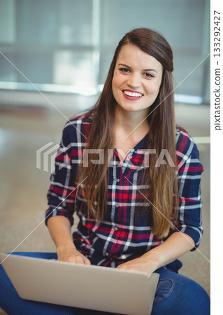 Woman wearing plaid shirt sitting on carpet with laptop, smiling at camera in office lounge 133292427