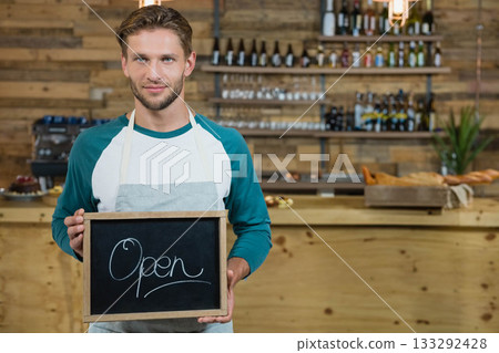 Male barista in teal shirt, apron standing at counter holding chalkboard Open sign beside pastries 133292428