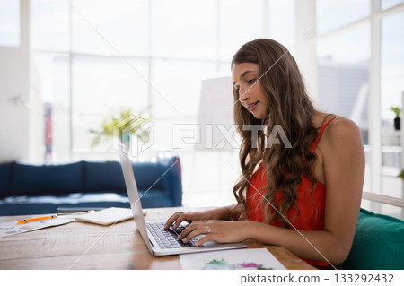 Female professional wearing red top typing on laptop at office table with charts, copy space Female professional wearing red top typing on laptop at office table with charts, copy space 133292432