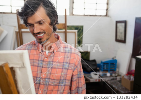 Mid-adult man painting on canvas at wooden easel in art studio with brushes and paint tubes 133292434