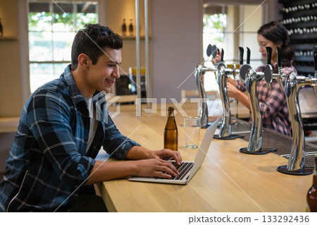 Male customer typing on laptop at bar with beer bottle, bartender operating draft taps, copy space Male customer typing on laptop at bar with beer bottle, bartender operating draft taps, copy space 133292436