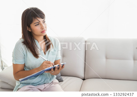 Asian woman in her twenties sitting on light sofa at home writing on clipboard, copy space Asian woman in her twenties sitting on light sofa at home writing on clipboard, copy space 133292437