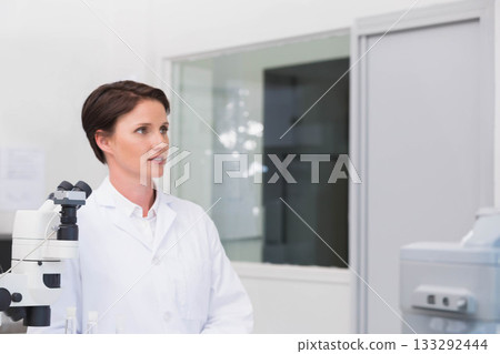 Female scientist wearing lab coat operating microscope in clinical lab beside diagnostic analyzer 133292444