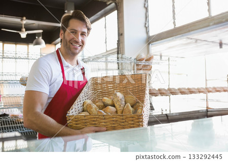 Male bakery worker wearing red apron smiling and holding basket of bread rolls behind glass counter 133292445