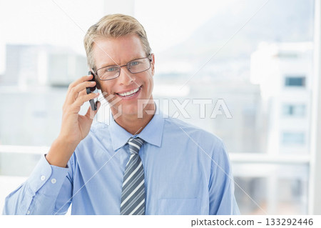 Businessman talking on smartphone at office window with city view, wearing glasses and striped tie 133292446