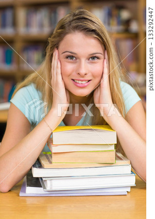 White teenager sitting at wooden table in library leaning on stack of textbooks by shelves smiling 133292447