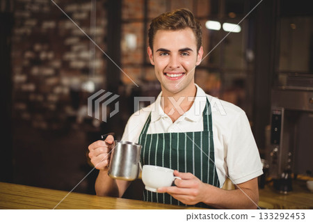 Smiling male barista pouring milk from pitcher into cup by espresso machine at coffee shop 133292453