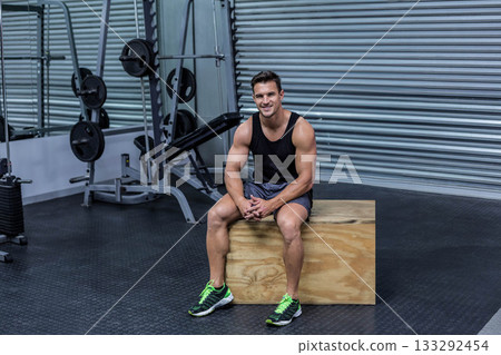 Male athlete in athletic wear resting on wooden plyometric box in gym near loaded squat rack 133292454