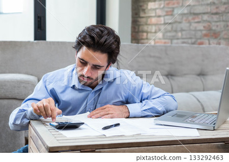 Man reviewing financial documents and using calculator at low wooden coffee table in living room Man reviewing financial documents and using calculator at low wooden coffee table in living room 133292463