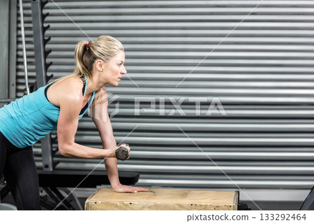 Woman in teal tank top, leaning over wooden box holding dumbbell at warehouse gym 133292464