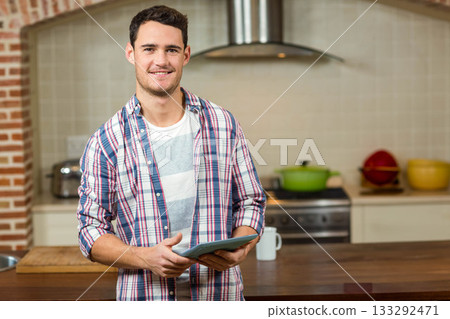 Man holding tablet and smiling while standing behind wooden island in home kitchen Man holding tablet and smiling while standing behind wooden island in home kitchen 133292471