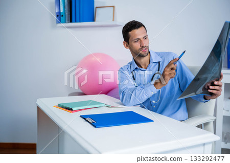 Mid adult male doctor examining X-ray film with stethoscope at desk in medical office, copy space 133292477