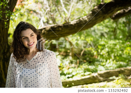 Woman wearing polka dot blouse leaning against moss-covered tree trunk in sunlit forest, copy space 133292492